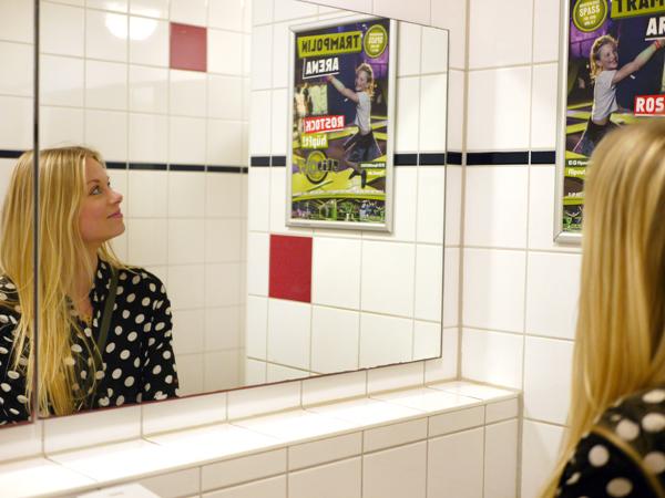Ein junge Frau schaut auf einen Plakatrahmen im Waschraum einer Toilette in der Gastronomie. Das Plakat wirbt für die Trampolin Arena in Rostock.